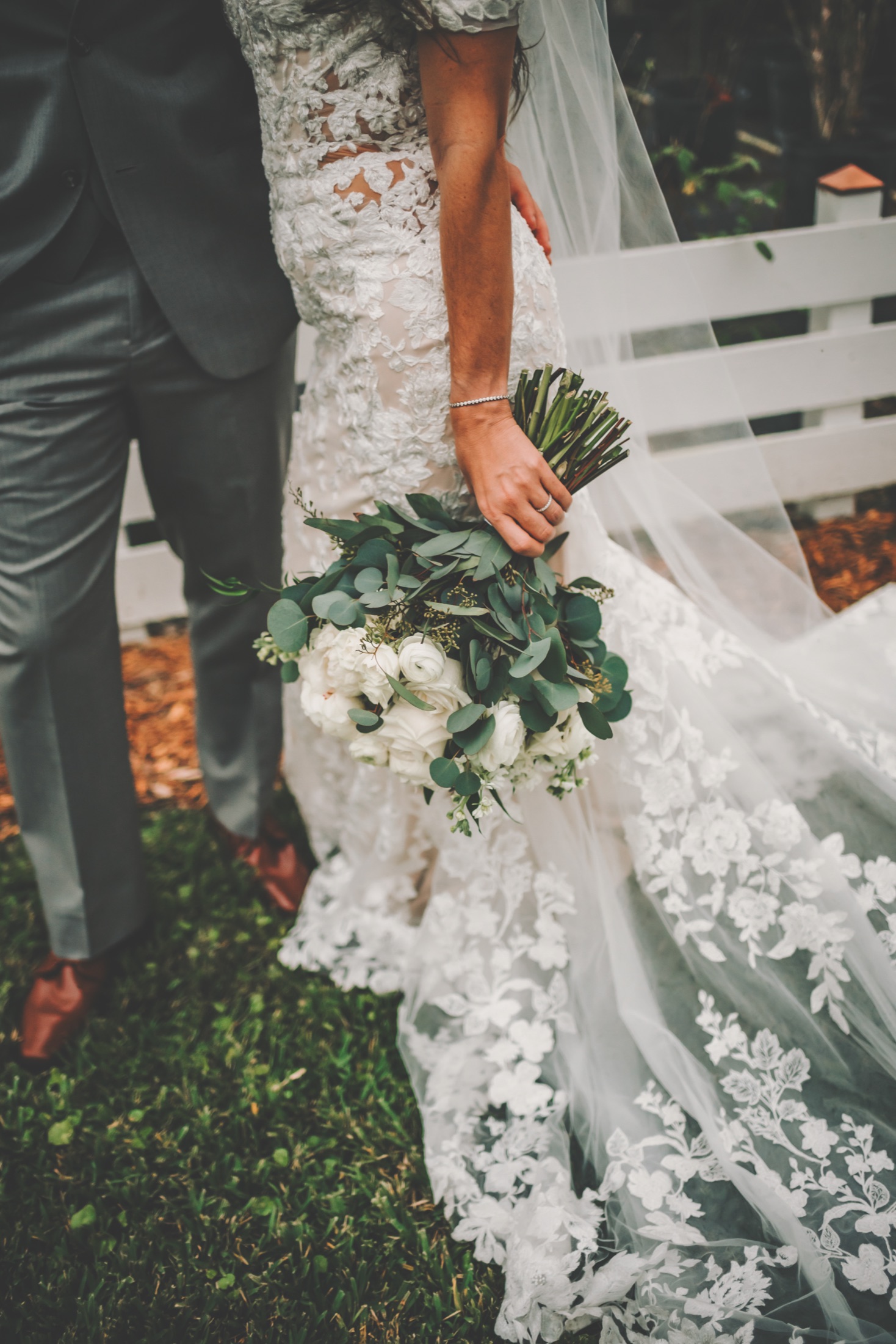 Bride holding a white-and-green bouquet beside her partner in The Arbor Estate gardens
