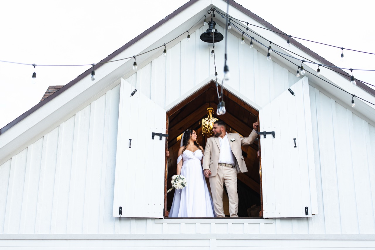 Kaila and Vincent holding hands in the open upper doorway of the white barn