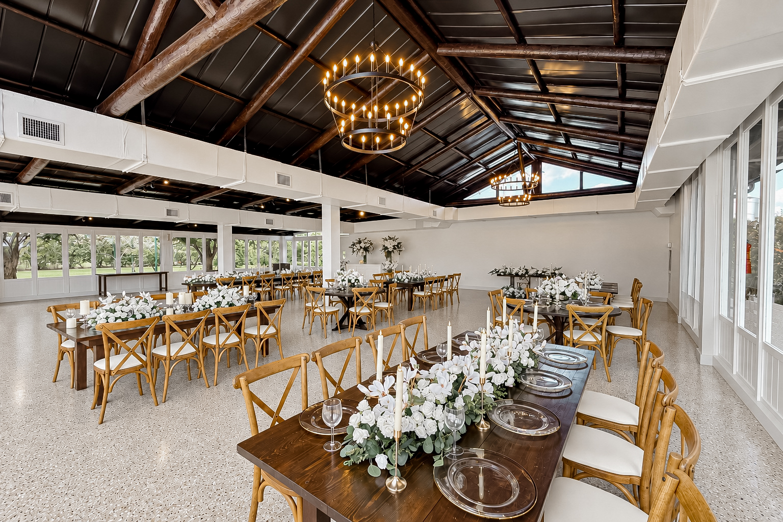 The Estate Room arranged with farmhouse tables, cross-back chairs, and white floral centerpieces beneath glowing chandeliers