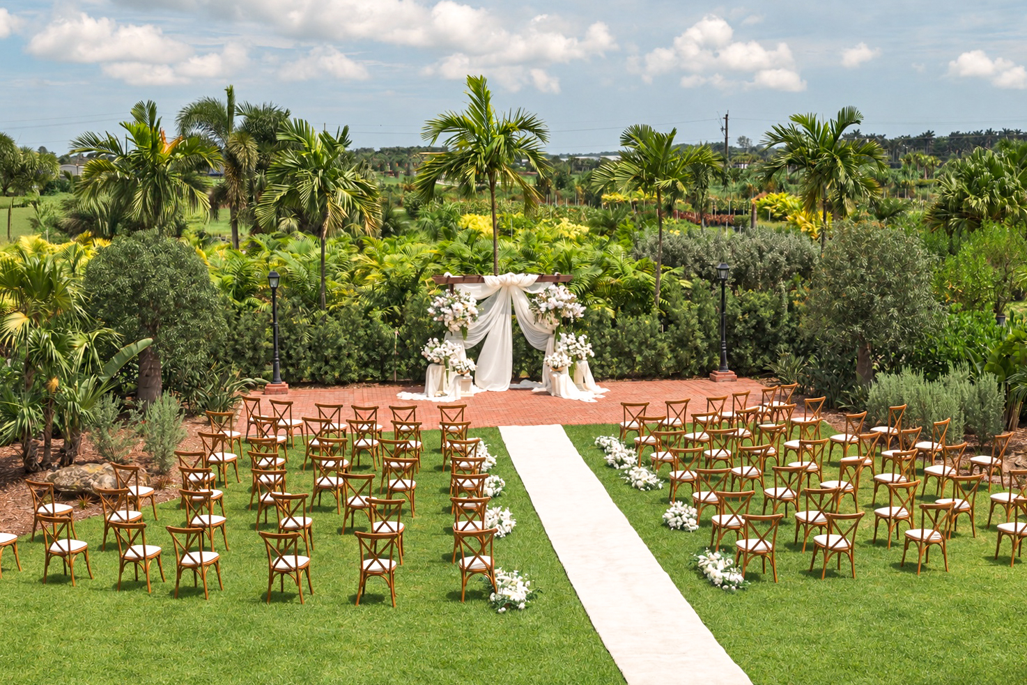 Garden ceremony setup at The Arbor Estate with cross-back chairs, a white aisle, and a floral arbor surrounded by tropical palms